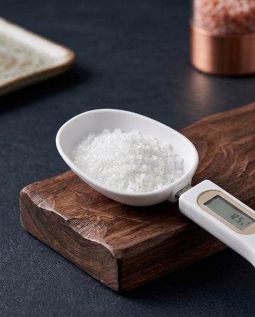 Digital measuring spoon scale holding coarse sea salt on a dark wooden board, display reading 12.5 g